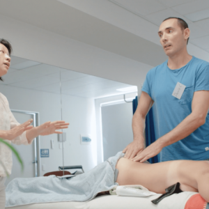 A therapist performs Bioelectric Meridian Therapy on a client as a colleague discusses BMT benefits in a sunlit clinic. Description: In a bright therapy room, one practitioner demonstrates Bioelectric Meridian Therapy whilst another explains its uses to an observer. Title: Discussing Bioelectric Meridian Therapy during treatment in a modern wellness clinic.