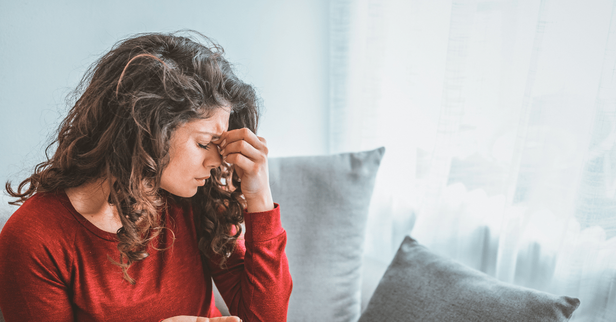 Bioelectric Meridian Therapy for Headache Relief Alt text: Woman in red shirt sits on sofa, pinching her nose from headache, with light streaming through nearby curtains. Description: Seeking relief from a headache, a woman sits under gentle daylight—Bioelectric Meridian Therapy may help restore comfort.