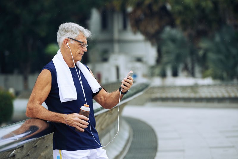 Senior man outdoors in sportswear checks BMT progress on phone Alt text: Older man with towel, sports bottle, and earphones reviews his Bioelectric Meridian Therapy updates on mobile by rail Description: An elderly athlete stands leaning on a curved railing, listening to music and exploring BMT benefits with his mobile device.